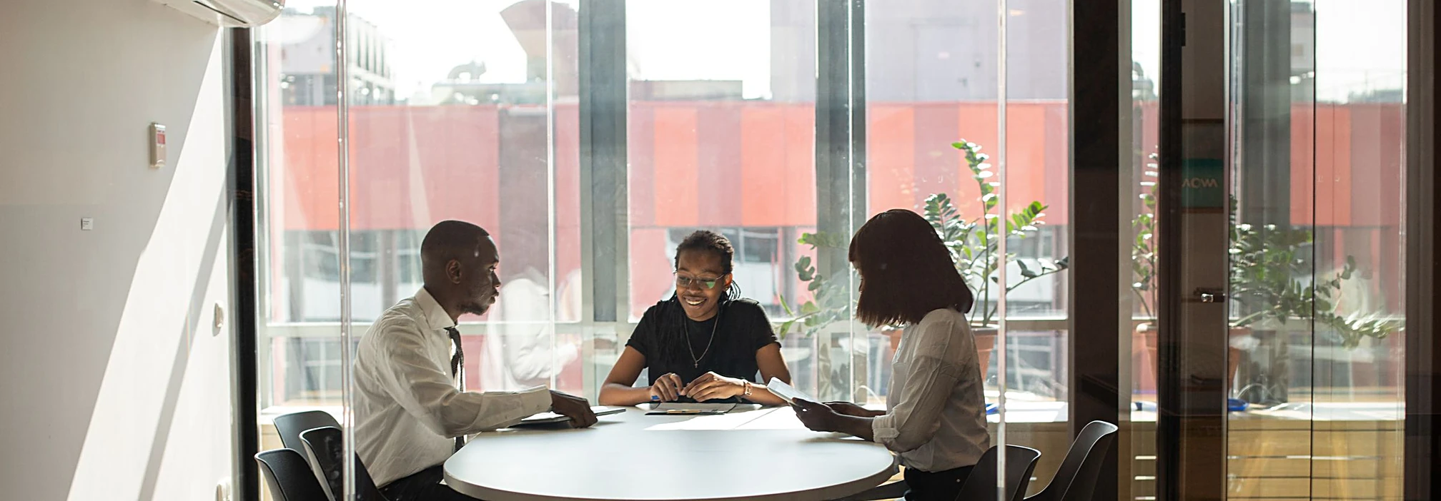 Three professionals collaborating around a round table in a sunlit glass boardroom
