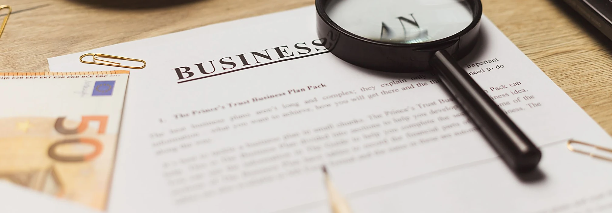 A business plan document under a magnifying glass, with coffee on a wooden desk